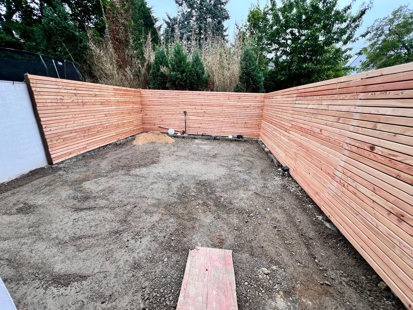 Vorher: Rohbau Terrassenbau Mettmann – Schotterbett und Holzzaun-Vorbereitung Vorher-Bild Terrassenbau Mettmann: Rohbau mit Schotterbett, Drainage und Holzzaun-Randabschluss